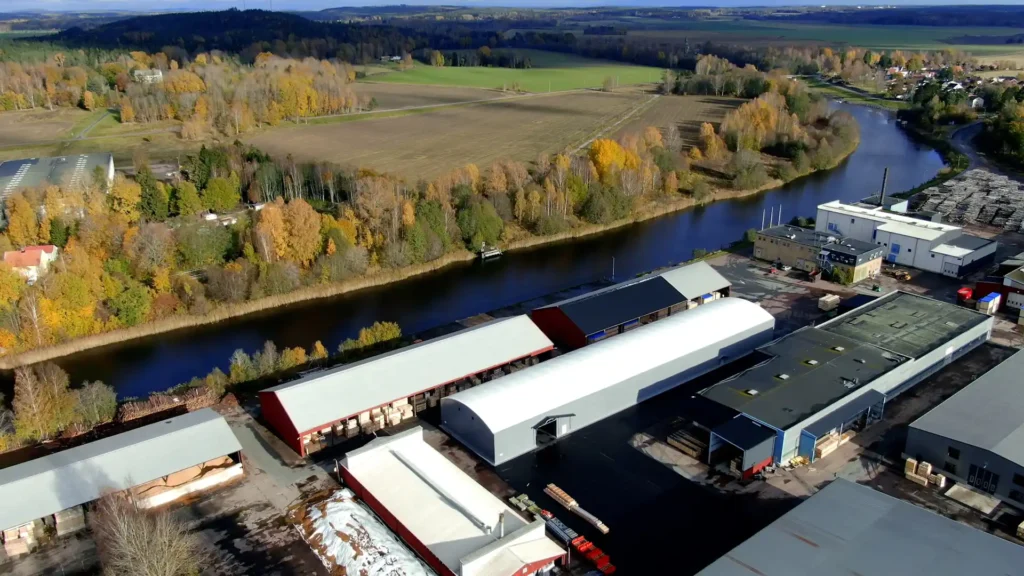 En grå hall från Hallbyggarna Jonsereds i fokus bland flera industribyggnader hos Wermland Logistikcenter med en å och åkermark i bakgrunden.