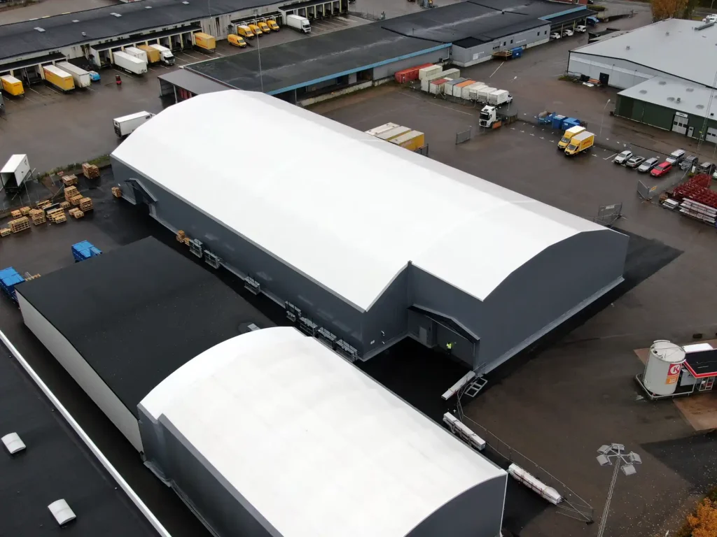 Två tälthallar med vita tak och grå dukar där den bortre och större hallen har ett specialkonstrurerat hörn för genomfart för lastbilar